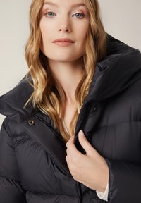 Czarna kurtka puchowa z wysokim kołnierzem, przednim zamkiem błyskawicznym i widocznym przeszyciem. Materiał wydaje się gładki i błyszczący, zapewniając izolację.