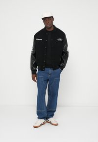 Fekete sportdzseki bőr ujjakkal, hímzett logókkal, fekete pólóval, kék farmernadrággal és fehér sportcipővel, fehér háttér előtt.