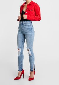 Červená džínová bunda, černý top, světle modré úzké džíny s rocehanými koleny a červené vysoké podpatky. Obsahuje rozparky na kotnících a třepící se okraje.