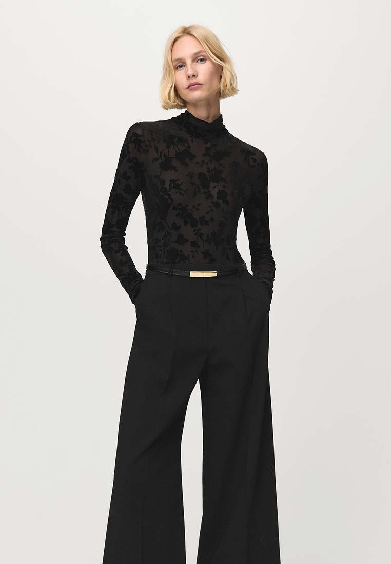 Femme aux cheveux blonds courts portant un haut noir transparent à motifs floraux à manches longues et un pantalon noir taille haute à jambes larges, les mains dans les poches.