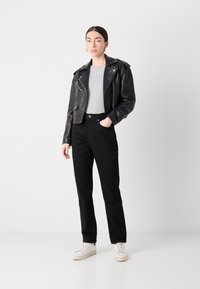 Giacca da biker nera in pelle con accessori in argento, t-shirt grigia aderente, jeans neri a vita alta e sneaker bianche. Posizione in piedi su uno sfondo neutro.