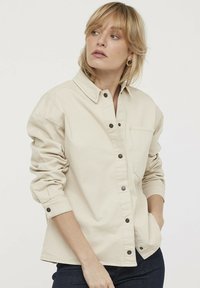 Camisa beige de botones confeccionada con una tela duradera, que presenta un cuello puntiagudo, mangas largas, un bolsillo en el pecho y detalles de botones de metal.