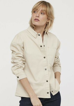 Chemise beige à boutons, fabriquée en tissu durable, dotée d'un col pointu, de manches longues, d'une poche poitrine et d'accents en métal sur les boutons.