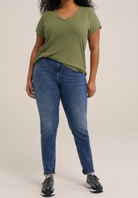 Olivgrön texturerad V-ringad topp i kombination med blå slim-fit jeans och grå sportskor. Har uppvikta ärmar och ett subtilt mönster.