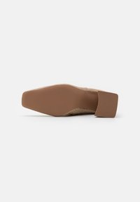 Chaussure à talon avec un dessus en cuir nude texturé présentant un talon carré, une semelle plate et un motif subtil inspiré des reptiles sur la surface.