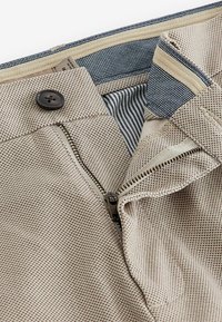 Tejido texturizado beige con cremallera, con un botón marrón y un forro interior a rayas. El material parece transpirable y duradero.
