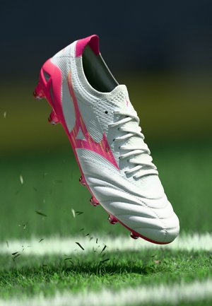 Crampon de football blanc et rose flottant au-dessus de l'herbe avec des brins dispersés et une ligne de terrain blanche en arrière-plan.