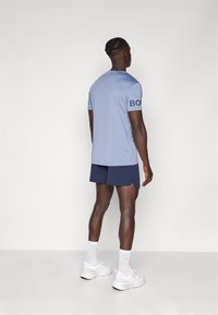 Camisa de manga curta azul-claro com o texto preto "BORN" na manga, combinada com calções azuis-marinho e sapatilhas brancas. Tecido suave, ajuste casual.