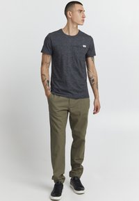 Marineblauwe gespoten crew-neck t-shirt met zakdetails, gecombineerd met beige chinos en zwarte lage sneakers. Tatoeages zichtbaar op de armen.