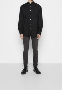Hombre de pie vistiendo una camisa negra de botón de manga larga, pantalones oscuros ajustados y zapatos negros de cordones, frente a un fondo liso.