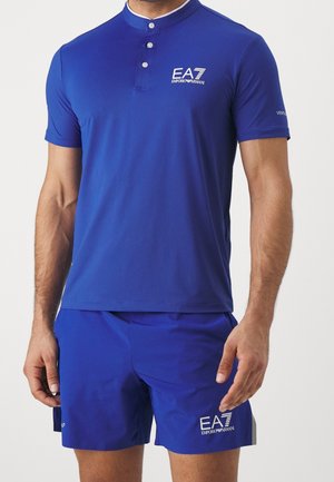 Mand iført en blå EA7 Emporio Armani kortærmet skjorte og matchende blå shorts med hvide logoer på bryst og lår.