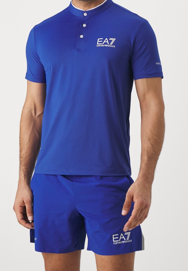 Homme portant une chemise à manches courtes bleue EA7 Emporio Armani et un short bleu assorti avec des logos blancs sur la poitrine et la cuisse.