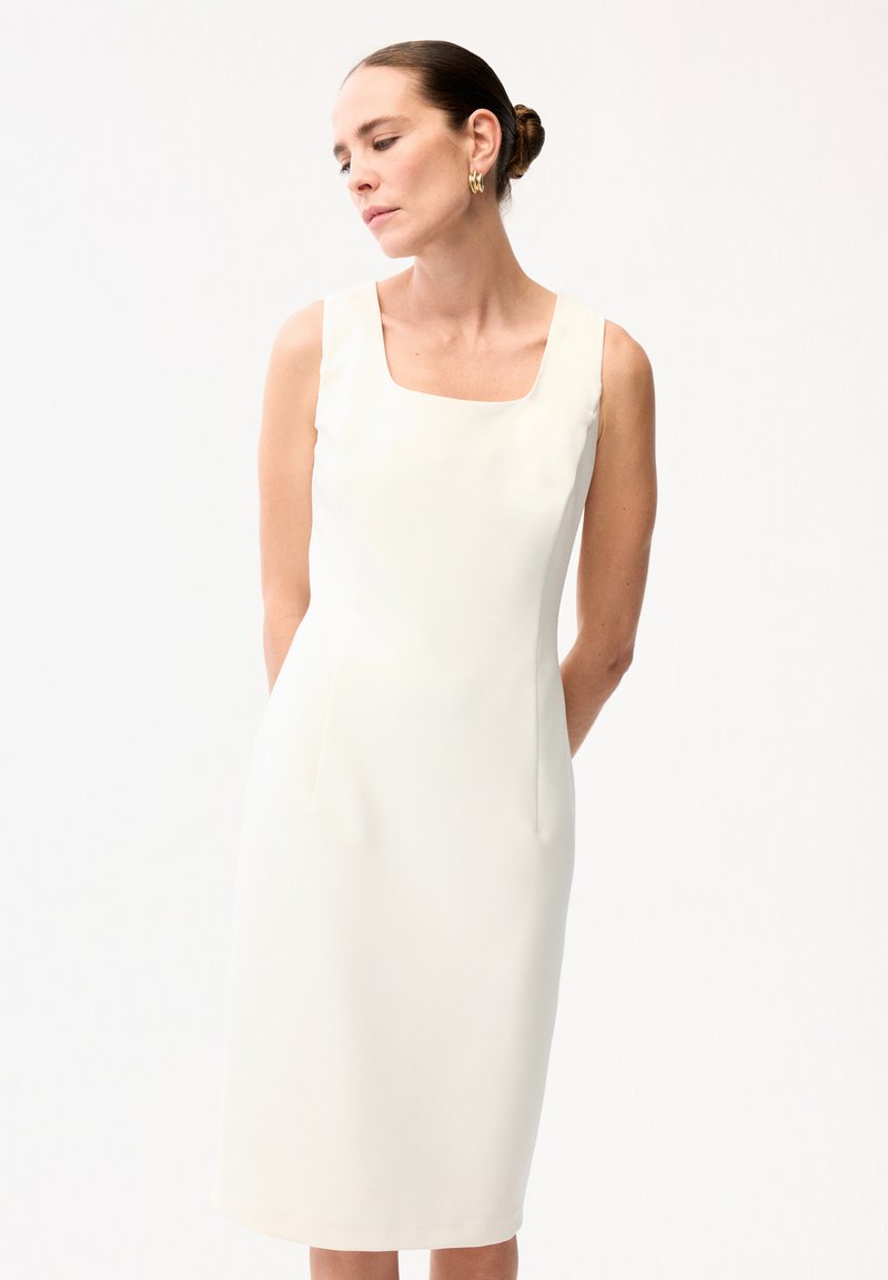 Femme aux cheveux attachés en chignon portant une robe midi blanche sans manches, ajustée, avec un décolleté carré et des boucles d'oreilles créoles dorées, les mains derrière le dos.