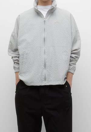 Personne portant une veste zippée gris clair texturée avec un col montant et un pantalon noir, mains dans les poches, debout devant un fond uni.
