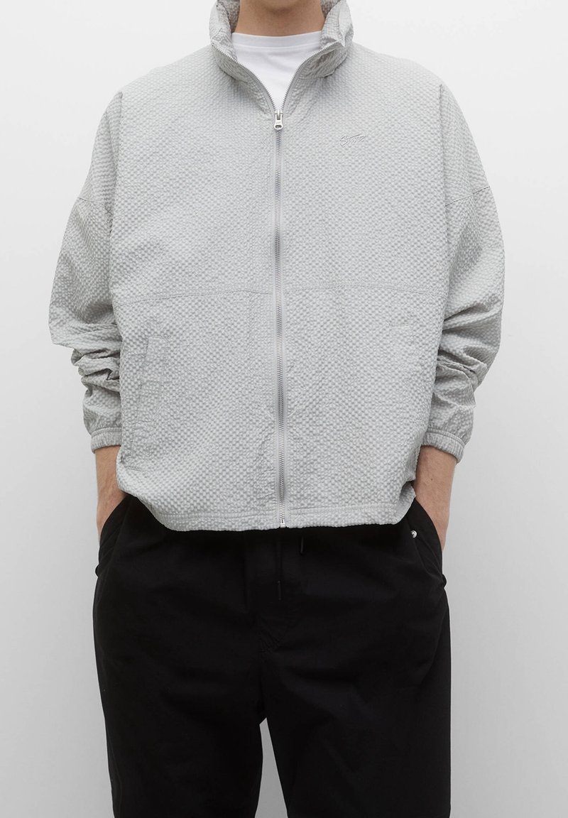 Personne portant une veste zippée gris clair texturée avec un col montant et un pantalon noir, mains dans les poches, debout devant un fond uni.