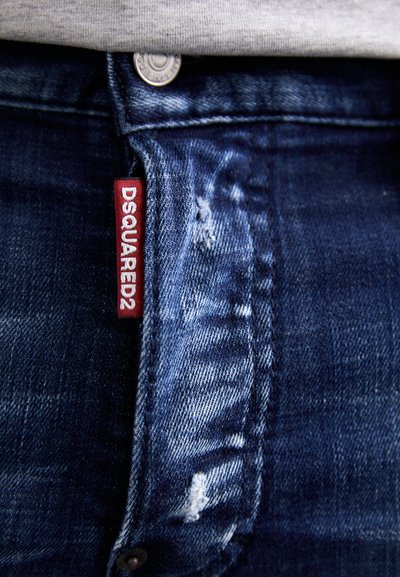 Tamno plave traperice s istrošenim izgledom, s vidljivom crvenom oznakom od tkanine na kojoj piše "DSQUARED2" i srebrnim detaljem na dugmetu.