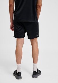 Homme portant un short de sport noir, un t-shirt noir, des chaussettes blanches et des chaussures de sport noires, debout, de dos sur un fond uni.