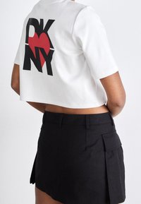 Bílé krátké tričko s velkým černým a červeným logem "DKNY" na zadní straně. Doplňeno černou sukní s kapsami.