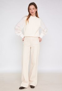 Pull sans manches crème en tricot avec un motif texturé, associé à un pantalon ample crème ; le mannequin se tient confiant avec les mains sur les hanches.