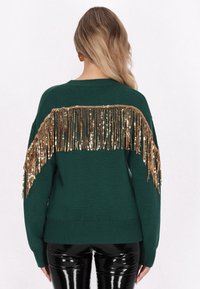 Pulover verde tricotat cu un fringă din paiete aurii pe spate, cu tiv elastic și mâneci lungi. Pantaloni negri lucioși completează ținuta.