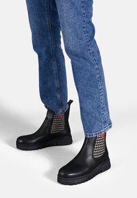 Stivale alla caviglia in pelle nera con pannelli laterali elastici a motivo, suole spesse e dettagli rossi. Indossati con jeans blu, che mettono in mostra il design dello stivale.