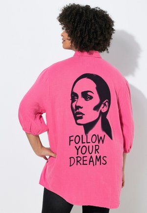 Kvinde med krøllet hår har en knaldrosa skjorte på med sort stencilportræt og teksten "FØLG DINE DRØMME" på ryggen.
