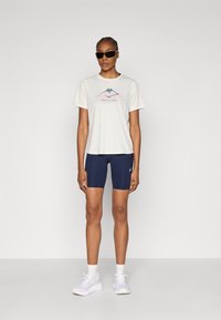 Ανοιχτό μπεζ μπλουζάκι με γραφικό βουνού, συνδυασμένο με σκούρα μπλε ποδηλατικά σορτς. Λευκά αθλητικά παπούτσια ολοκληρώνουν την εμφάνιση, κατάλληλη για αθλητική ένδυση.