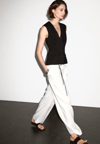 Gilet nero senza maniche con scollo a V e chiusura con bottoni, abbinato a pantaloni bianchi larghi con coulisse. Materiali lisci e linee pulite.