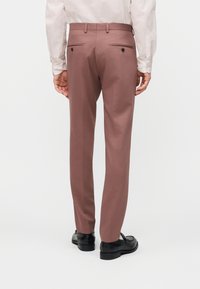 Άνδρας που φοράει παντελόνι φόρμας σε χρώμα ροζ σκόνης και μαύρα παπούτσια, εμφανιζόμενος από πίσω σε απλό λευκό φόντο.