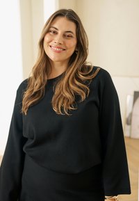 Black, long-sleeve sweater with a relaxed fit. Soft texture, round neckline, and loose cuffs. Warm tone, worn by a smiling woman in a bright setting.
