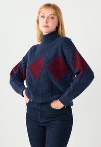 Maglione lavorato a maglia blu e bordeaux con collo alto, caratterizzato da motivi a diamante e tessuto a rilievo. Abbinato a jeans di denim scuro.