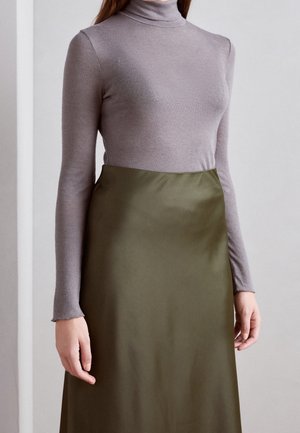 Femme portant un pull-col roulé violet clair et une jupe taille haute vert olive, debout devant un fond clair uni.