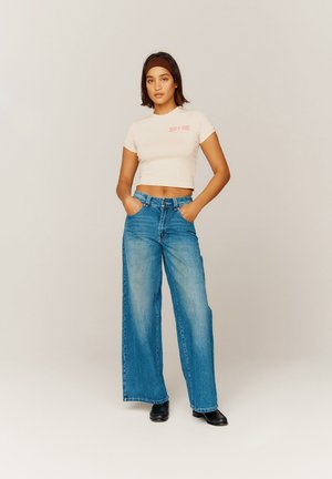 Helles beiges, kurzes T-Shirt mit dem Schriftzug "HOUSE OF SUNNY", kombiniert mit weit geschnittenen blauen Jeans. Das Outfit hat eine lockere Passform und einen leichten Verblassen.