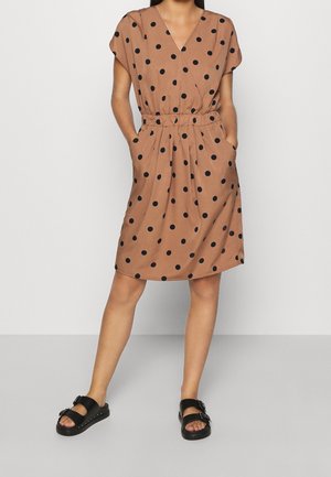 Robe marron à pois avec un décolleté en V, des manches courtes, une taille élastique, des poches latérales et un tissu fluide. Portée avec des sandales noires.