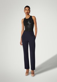 Sort ærmeløs bodysuit med blomstertekstur, kombineret med marineblå skræddersyede bukser med lommer, suppleret med beige højhælede sko.