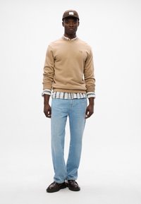 Felpa beige con scollo rotondo, indossata sopra una camicia bianca e grigia a righe. Jeans in denim azzurro chiaro e cappellino marrone, abbinati a scarpe marroni.