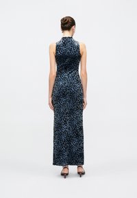 Donna con i capelli raccolti in uno chignon che indossa un abito lungo fino al pavimento senza maniche con stampa leopardata blu e nera e sandali neri con tacco basso, vista di schiena.