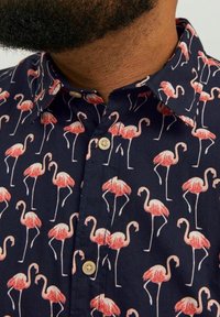 Camisa de manga corta hecha de tejido azul marino con un patrón repetido de flamencos rosas, con un cuello clásico y botones beige.