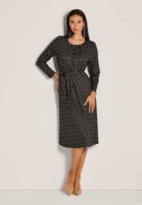 Frau, die ein schwarzes Midi-Kleid mit Kettenmuster, langen Ärmeln und einem gebundenen Taillengurt trägt, kombiniert mit beigen spitzen Absätzen und langen Ohrringen.