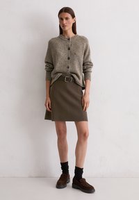 Frau in einem beigen, geknöpften Pullover, der in einen braunen, mit Gürtel versehenen Rock gesteckt ist, kombiniert mit schwarzen Socken und dunkelbraunen Slippers, die vor einer weißen Wand steht.