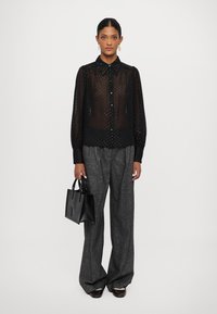Transparante zwarte blouse met knoopsluiting en een stippenpatroon, lange pofferige mouwen en een zwarte kraag, gecombineerd met grijze wijde broek en een kleine zwarte tas.