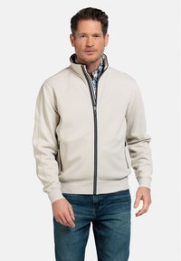 Lichtbeige zip-up jack met zwarte accenten. Beschikt over een hoge kraag en twee zijzakken. Draag het over een gestreept shirt, gecombineerd met blauwe jeans.