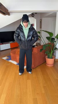 Juoda pūkinė striukė blizgiu paviršiumi, derinama su žalia džemperiu su gobtuvu, plačiomis juodomis kelnėmis ir sidabriniais batais, fone šviesiai apšviestoje patalpoje.