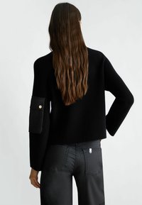 Maglione nero a coste con design cropped e maniche lunghe. Presenta una tasca laterale con chiusura a bottone. Indossato con pantaloni neri.