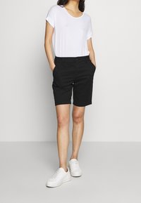 Černé šortky s přiléhavým střihem a bočními kapsami, v kombinaci s bílým tričkem s krátkým rukávem a bílými teniskami.