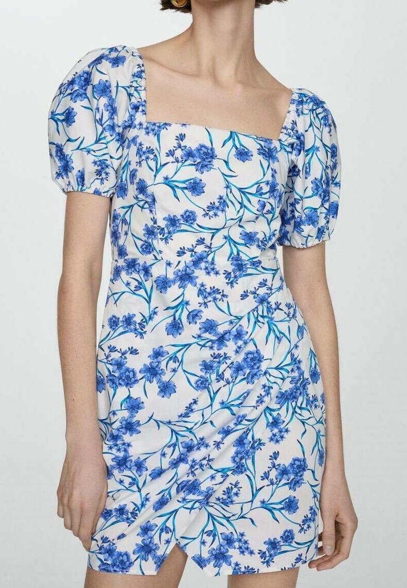 Femme en robe blanche avec imprimé floral bleu et manches courtes bouffantes, debout sur un fond uni.