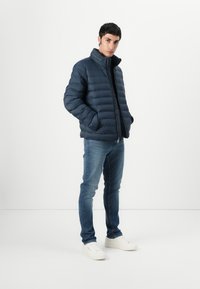 Navy padded jacket with a high collar, zip closure, and side pockets, paired with blue jeans and white sneakers. Smooth texture.