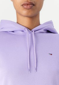 Sudadera con capucha morada con cordones. El material parece suave; cuenta con un pequeño logo en rojo, blanco y azul en la zona del pecho.