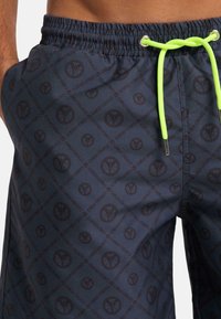 Dark blue swim shorts with a repeating black logo pattern, elastic waistband, and neon green drawstring for adjustment. Lightweight fabric.