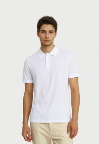 Nuori mies, joka on pukeutunut valkoiseen lyhythihaiseen polo-paitaan ja beigeihin housuihin, seisoo yksinkertaista valkoista taustaa vasten.